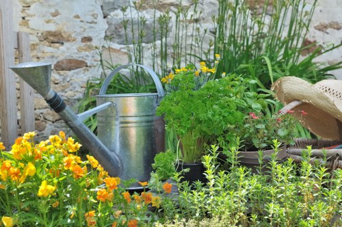 The image depicts a vibrant garden scene featuring a variety of plants and garden elements. In the foreground, there are bright yellow flowers with orange blooms, contrasting with lush green foliage. Behind the flowers, a weathered stone wall provides a rustic backdrop. To the right, a metal watering can is placed among potted plants, including leafy green herbs and flowering plants with small pink blossoms. Tall, grass-like plants with narrow leaves grow behind the watering can, adding height and texture to the garden bed. On the far right, a straw garden hat rests atop the soil or a container, suggesting outdoor gardening activity. The overall scene showcases a well-tended, diverse garden space that highlights the use of various plant types, garden tools, and containers, aligned with professional gardening and landscaping services in the Clapham area. The weather appears bright and sunny, enhancing the vibrant colours of the garden.
