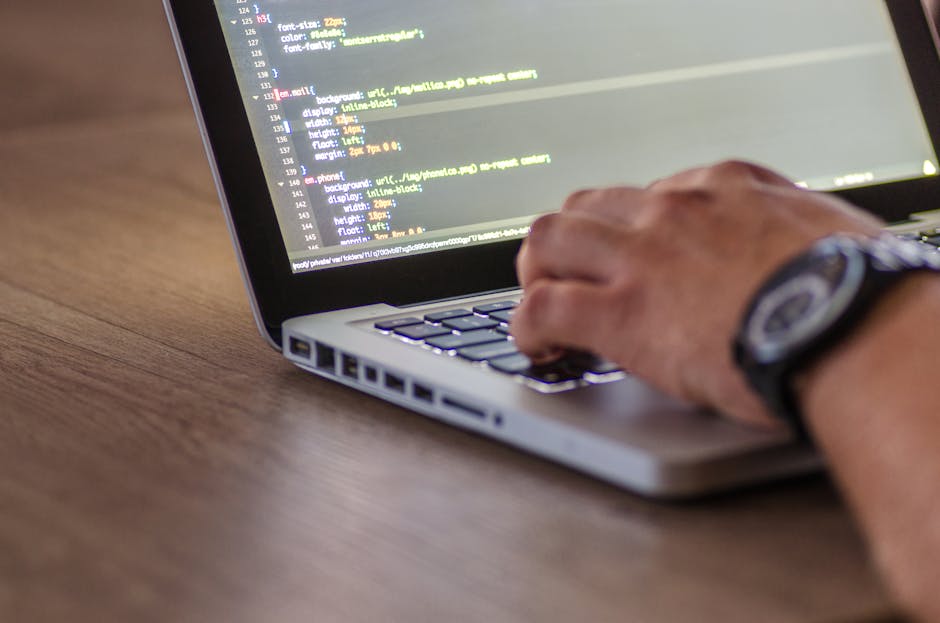 A close-up view of a person's hand typing on a silver laptop keyboard placed on a wooden surface. The laptop screen displays lines of code with syntax highlighting in various colors. The person's wris